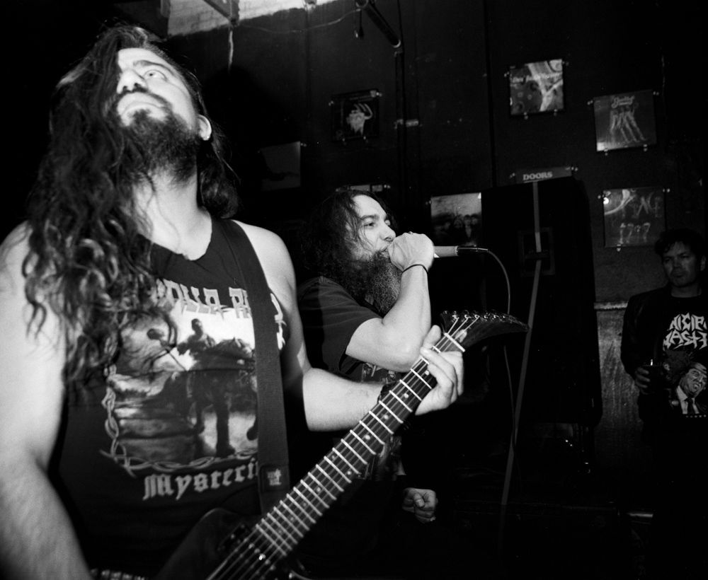 Black-and-white film photo of a heavy metal band performing onstage, with a guitarist in the foreground and vocalist behind him. Developed and printed in a traditional wet darkroom by the photographer, Madison Avery Somerville.