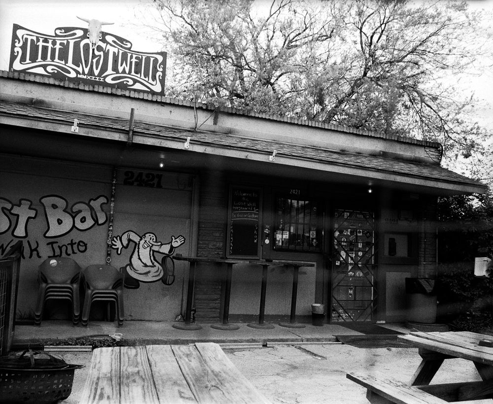 Black-and-white view of a rustic bar storefront with signs, porch, and picnic table. Shot on medium format film by Madison Avery Somerville. Developed and printed in a traditional wet darkroom by the photographer.