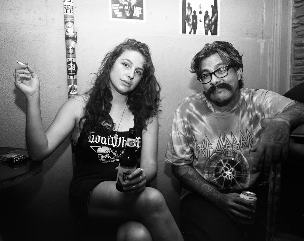 Two people sitting in a dim room, holding drinks, one smoking, with posters on the wall. Shot on medium format film by Madison Avery Somerville. Developed and printed in a traditional wet darkroom by the photographer.