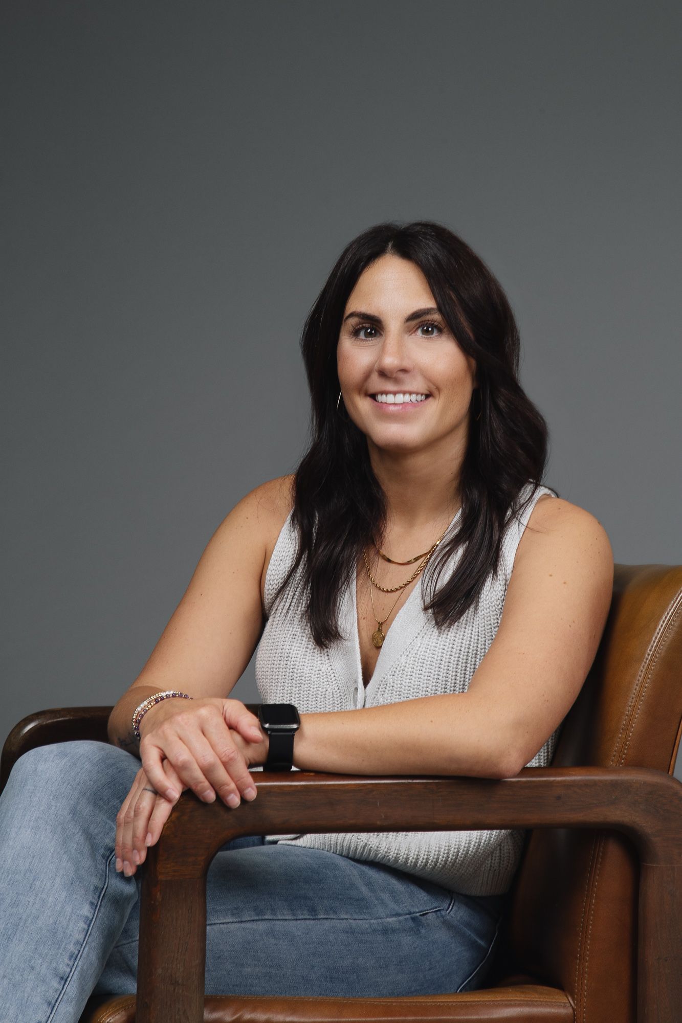 A smiling woman with dark hair sits in a leather chair, wearing a light-colored sleeveless top and a wristwatch.