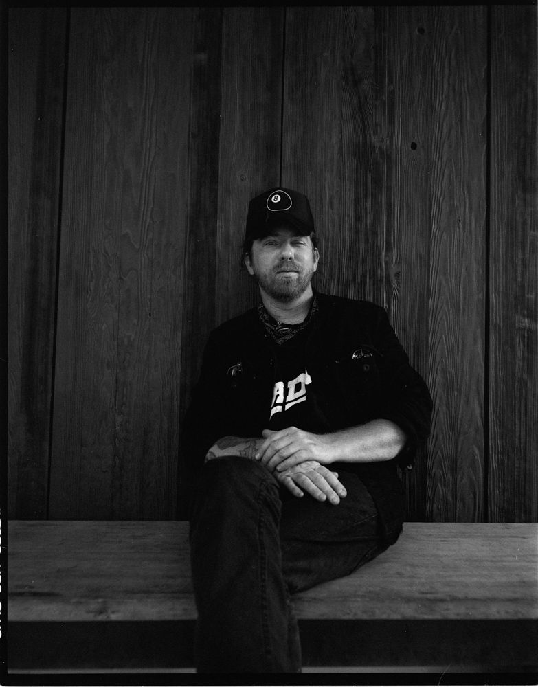 Black-and-white portrait of a seated man in a cap against a wooden wall. Shot on medium format film by Madison Avery Somerville. Developed and printed in a traditional wet darkroom by the photographer.