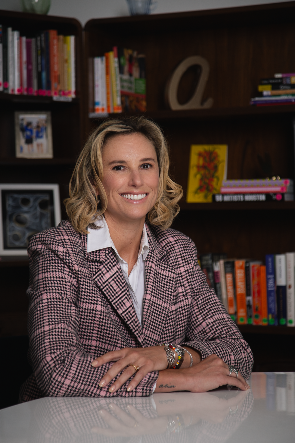 Woman seated at a desk in front of bookshelves, wearing a patterned blazer and smiling