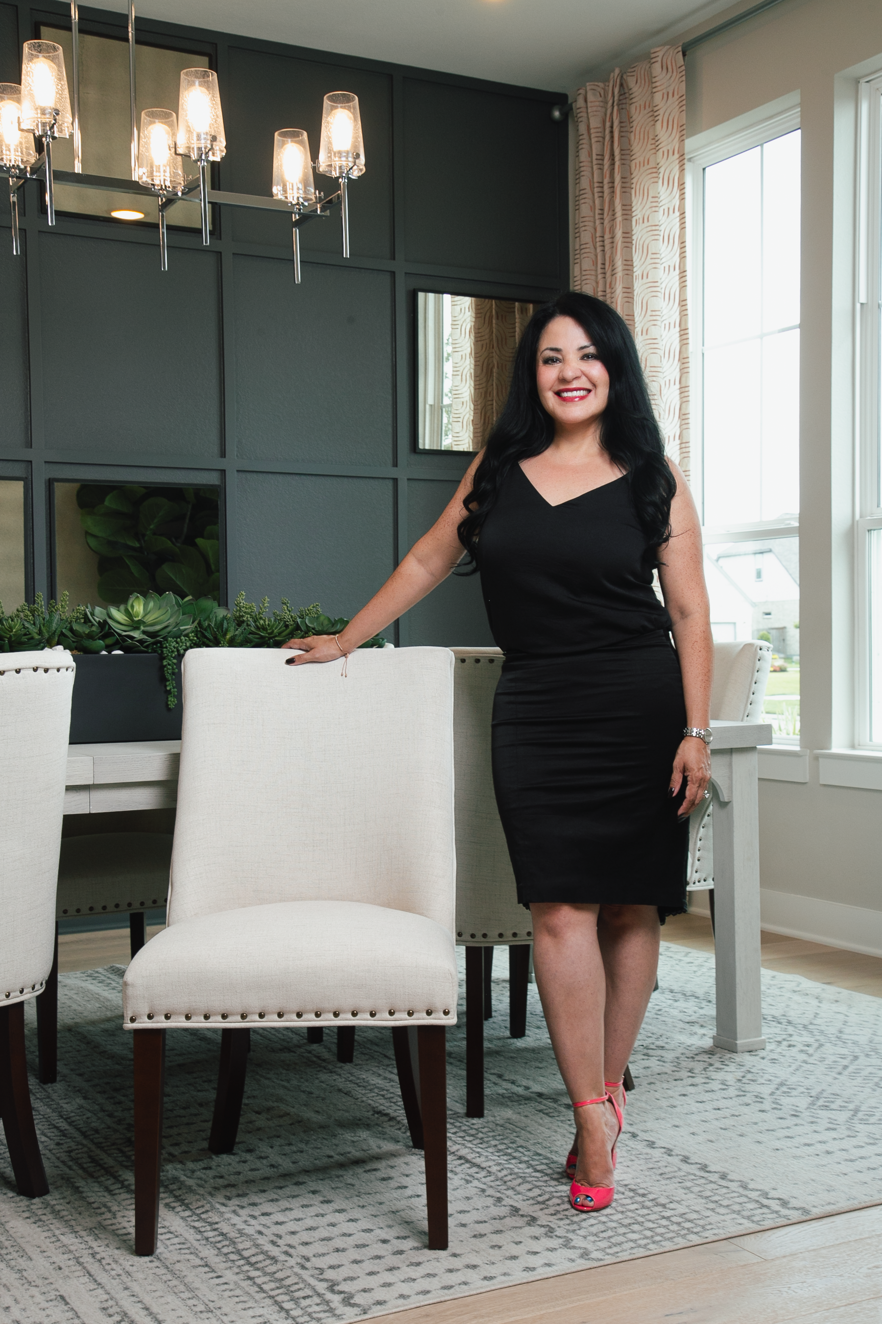 Woman in black dress leans on chair in dining room. Pink heels.