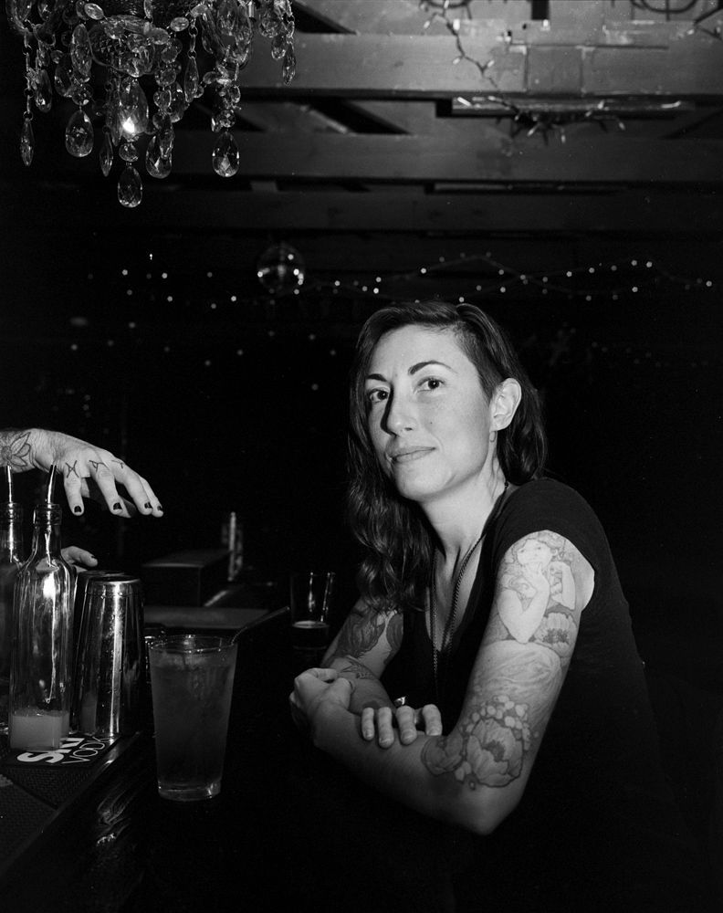 Woman with tattooed arms seated at a dim bar, looking at the camera under hanging lights. Shot on medium format film by Madison Avery Somerville. Developed and printed in a traditional wet darkroom by the photographer.