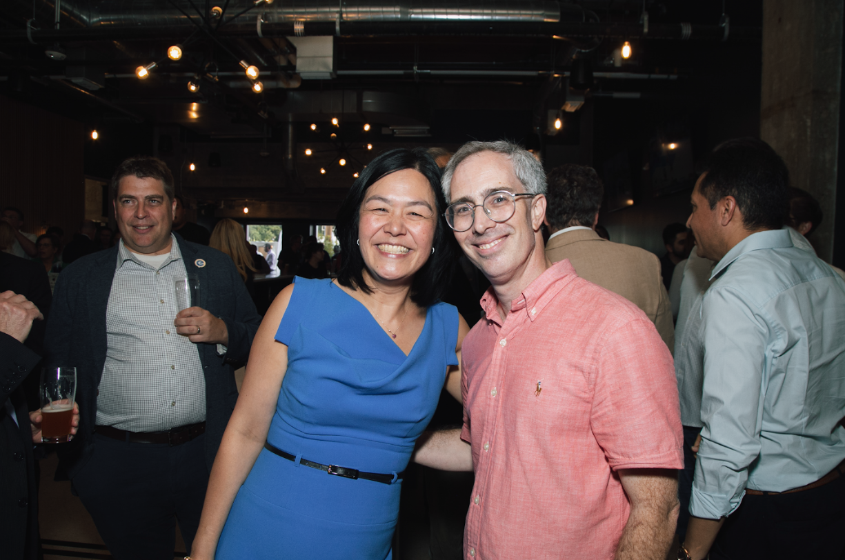 A smiling woman in a blue dress and a man in a pink shirt pose for a photo at a social gathering.
