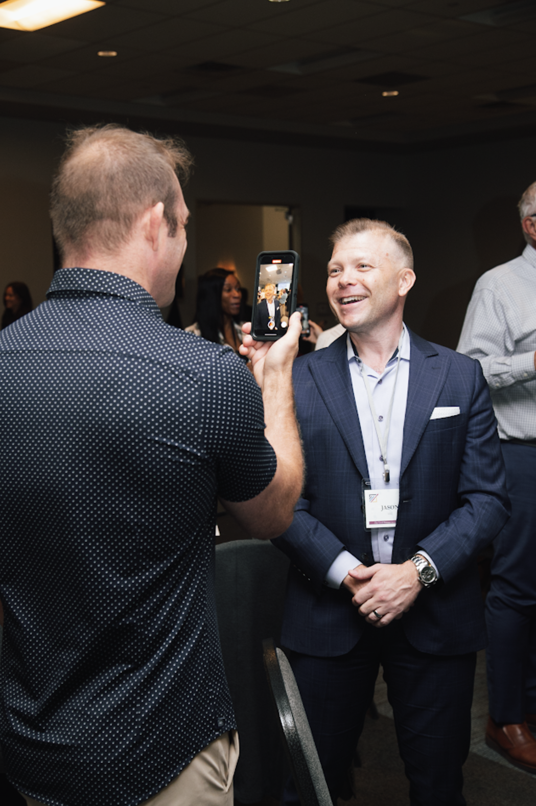 A man in a patterned shirt films a man in a navy blue suit who is smiling during an indoor professional event.