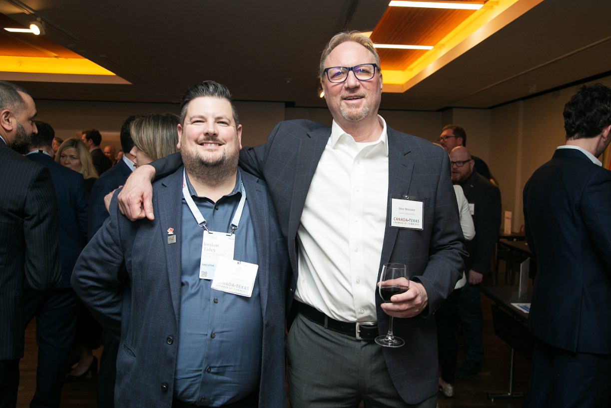 Two men in formal attire pose for a photo at an indoor event; the one on the right holds a glass of red wine.