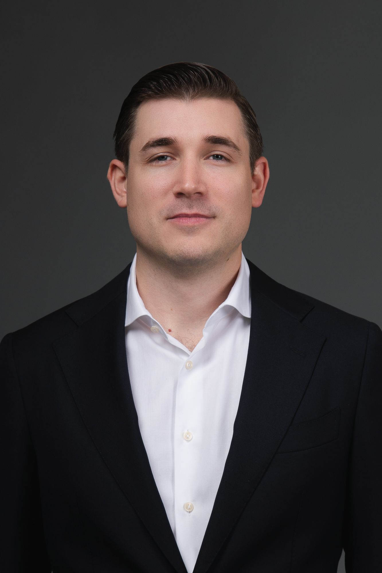 A person in a dark blazer and white button-down shirt against a plain gray background.