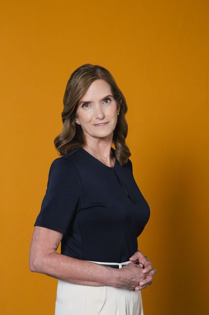 Brand photograph of a woman standing in a navy dress top and white pants against a bright yellow background