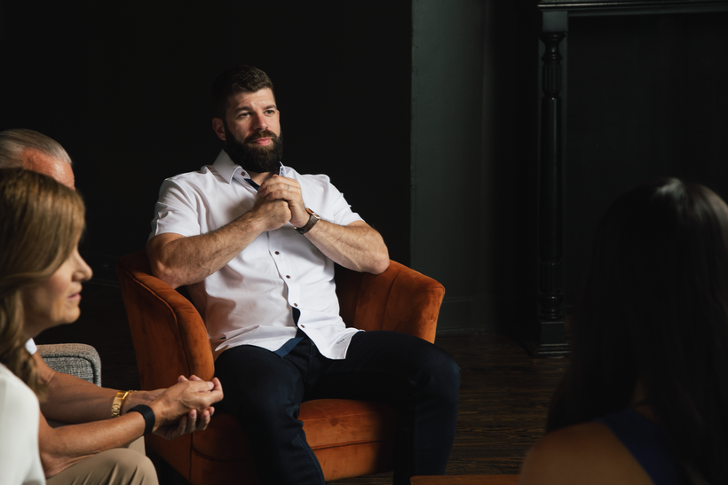 A man sits on a chair with his hands clasped in front of him. He sits with two other people and listens to another person across the room.