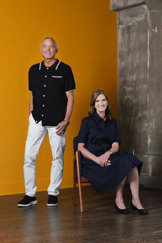 Brand photograph of two people posing against a mustard-yellow wall, one standing and one seated on a chair in a dark dress.