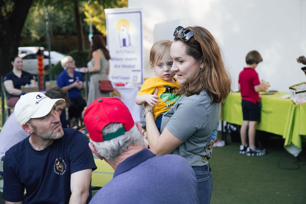 Woman holding a child at an outdoor community event, with people and green tables in the background