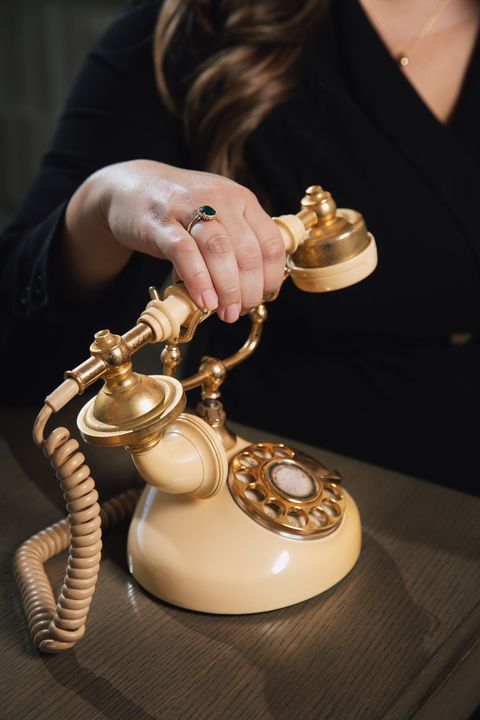 A person wearing a ring holds the handset of a vintage, cream-colored rotary telephone on a dark surface.