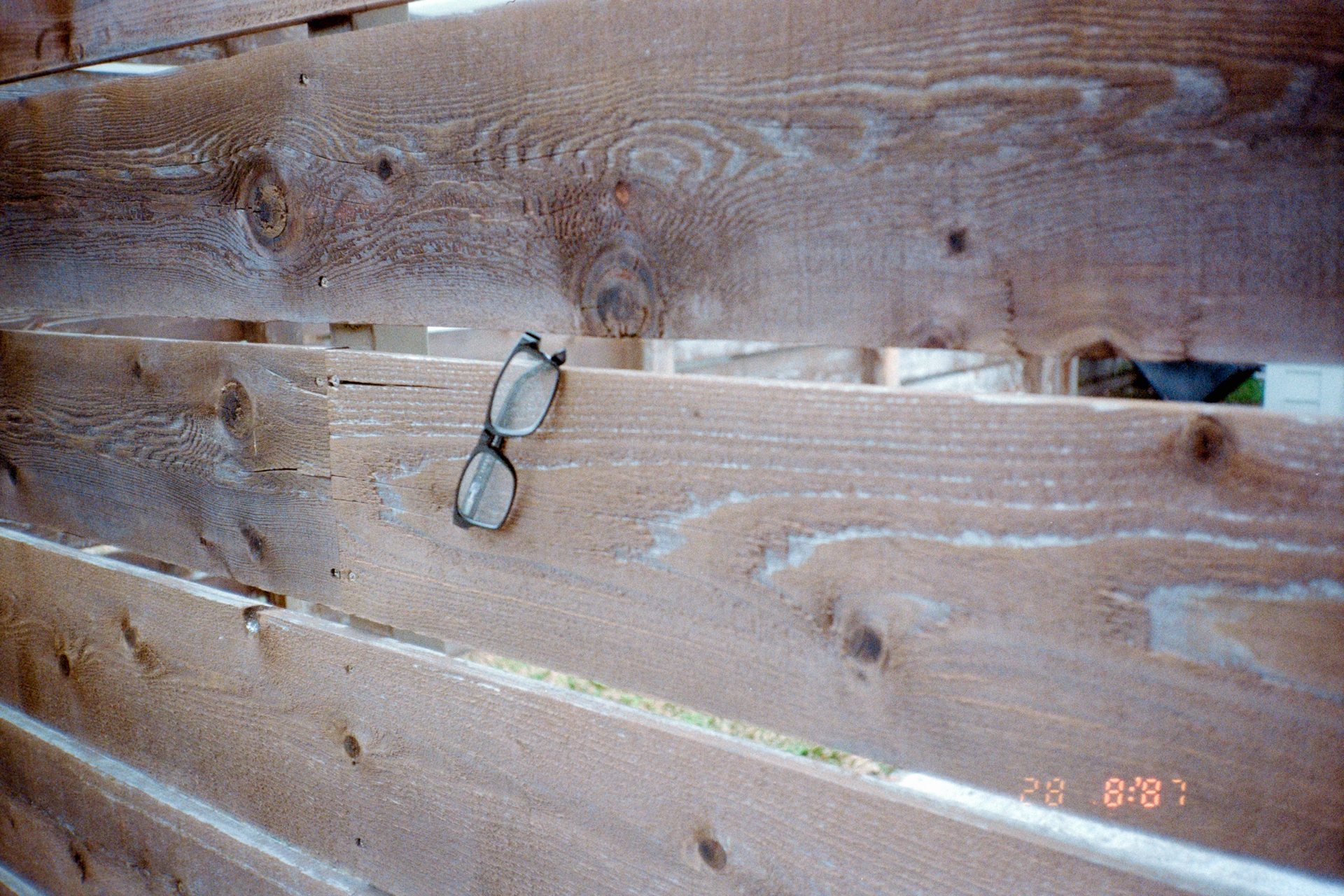 A pair of black-rimmed eyeglasses hung on a horizontal wooden fence plank.
