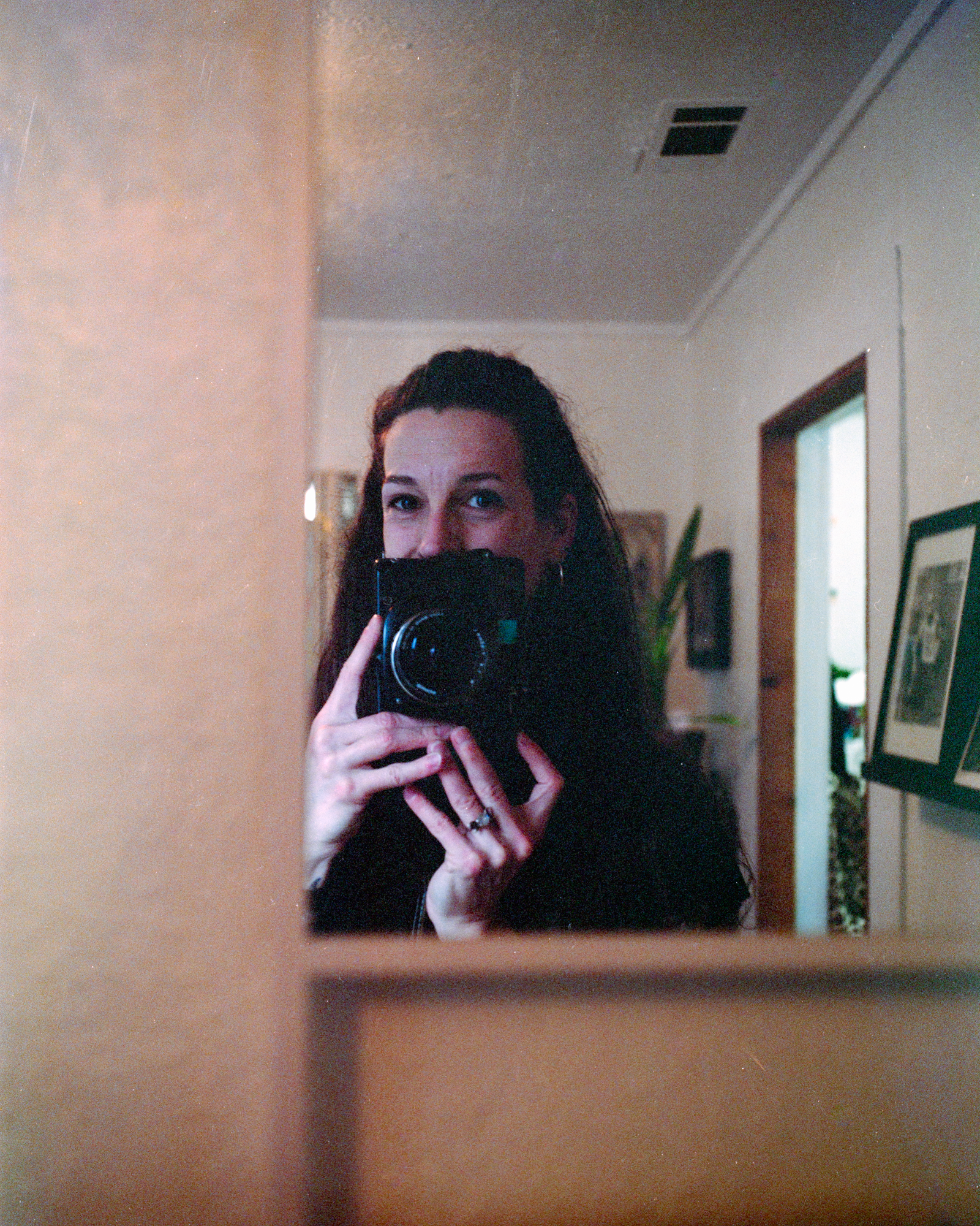 A person with long dark hair holds a camera up to their face, capturing a reflection in a mirror inside a room.