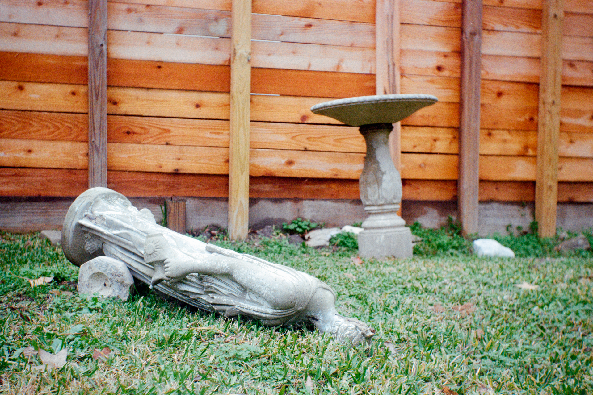 A broken stone column lies face down in a grassy yard near a standing birdbath in front of a wooden fence.