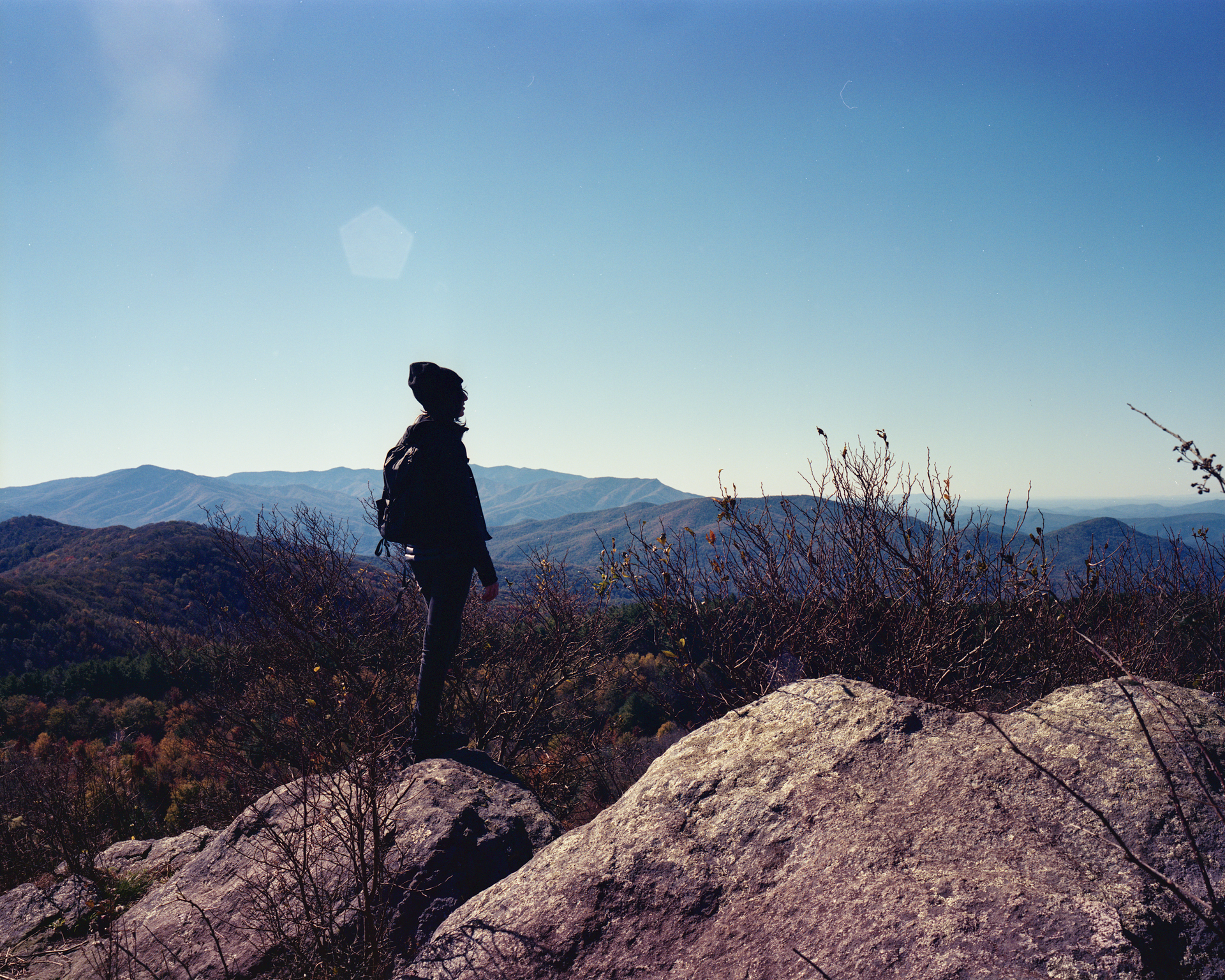 A silhouette of a hiker standing on a rocky mountain peak, overlooking a vast, hilly landscape under a clear blue sky.