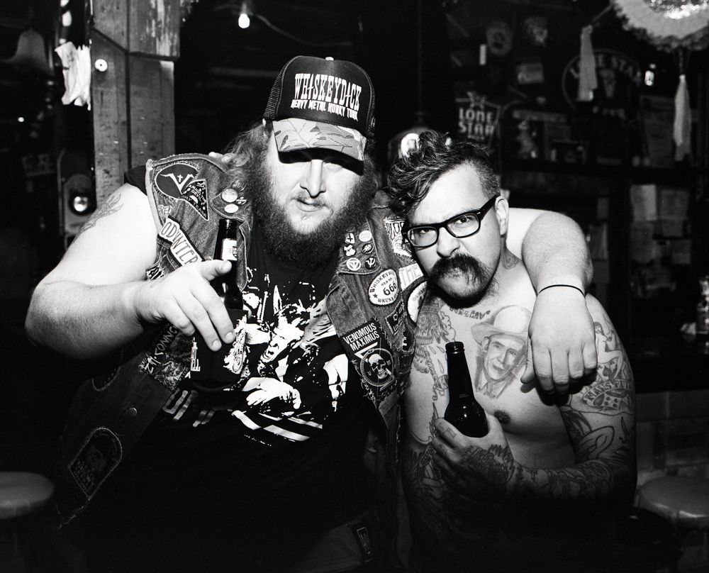 Two men posing in a dim bar, one holding a bottle and both wearing caps and glasses. Shot on medium format film by Madison Avery Somerville. Developed and printed in a traditional wet darkroom by the photographer.