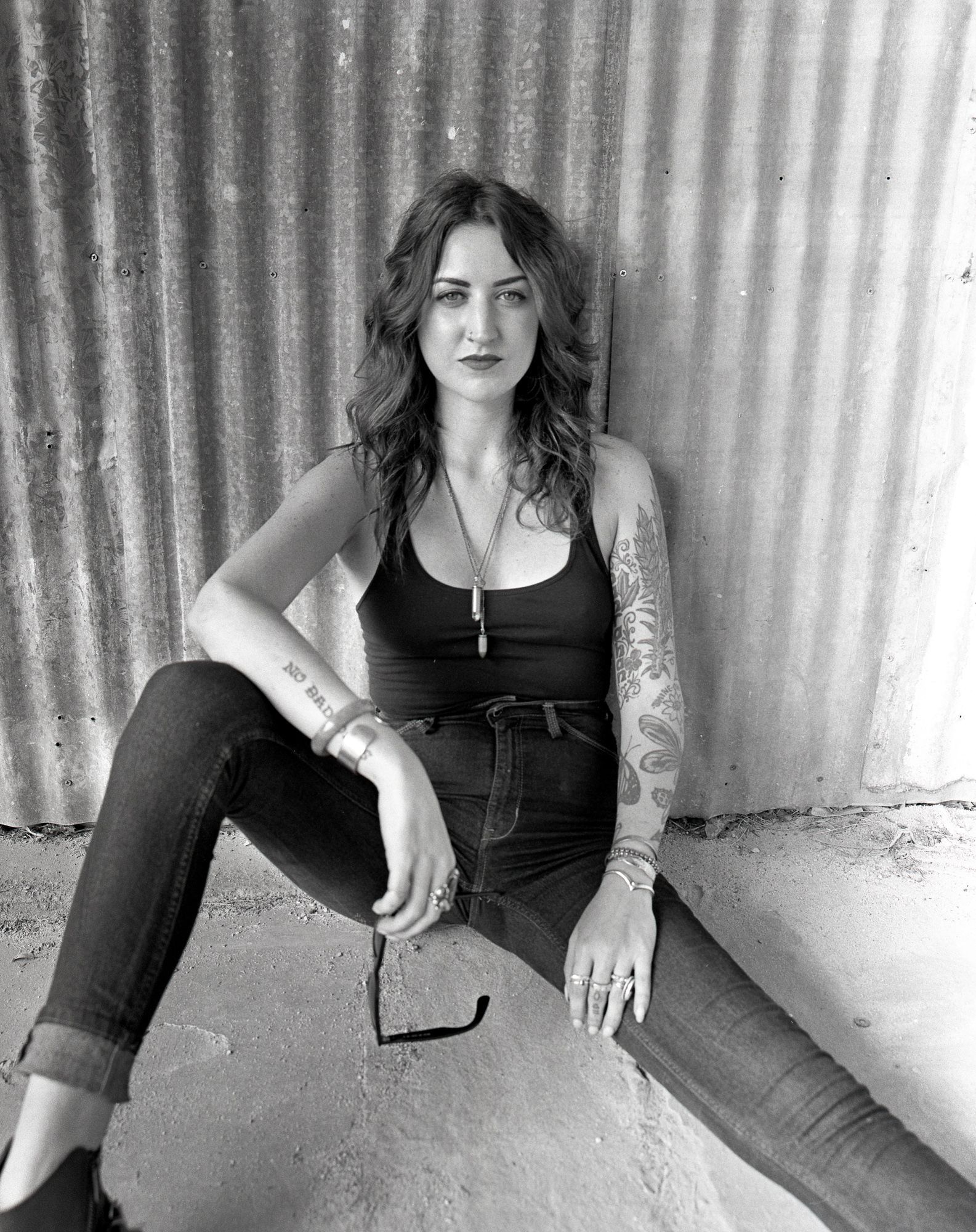 Person sitting on the floor against a corrugated wall, wearing black top and jeans, holding sunglasses. Shot on medium format film by Madison Avery Somerville. Developed and printed in a traditional wet darkroom by the photographer.
