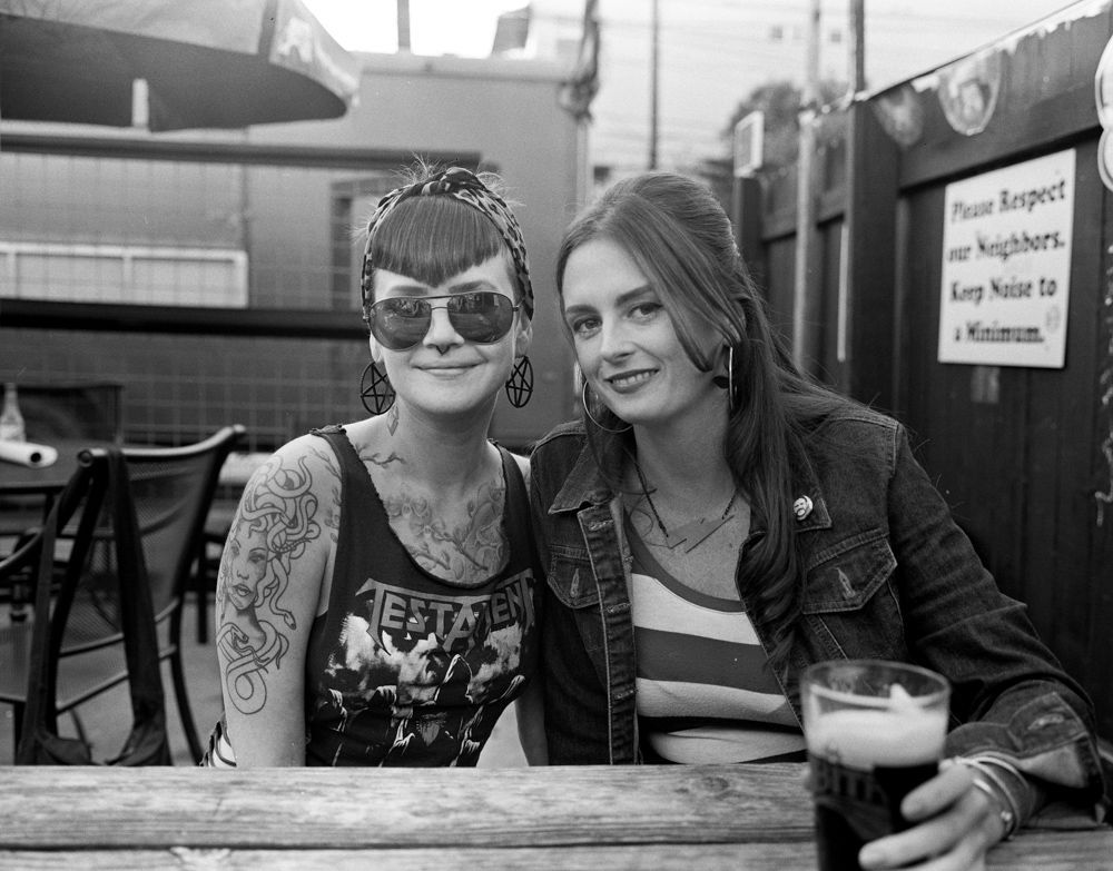Two women smiling at an outdoor table, one holding a pint, in a casual patio setting. Shot on medium format film by Madison Avery Somerville. Developed and printed in a traditional wet darkroom by the photographer.