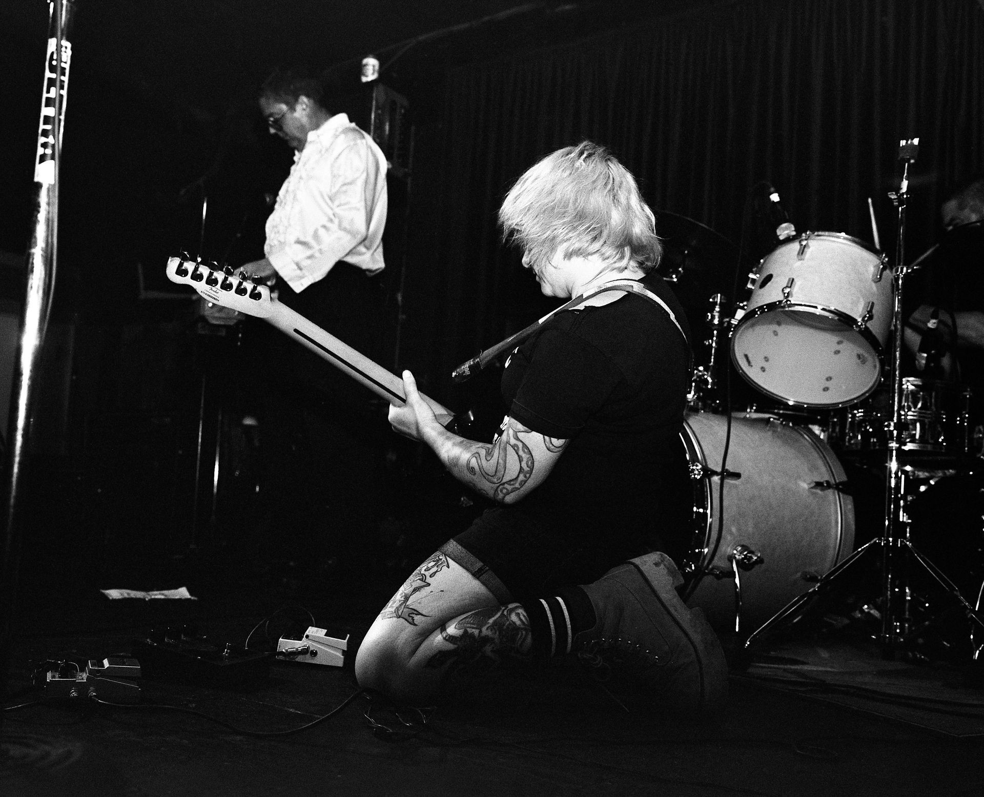 Black-and-white film photo of a kneeling guitarist onstage, with a drummer and another musician in the background. Shot on medium format film. Developed and printed in a traditional wet darkroom by the photographer, Madison Avery Somerville.