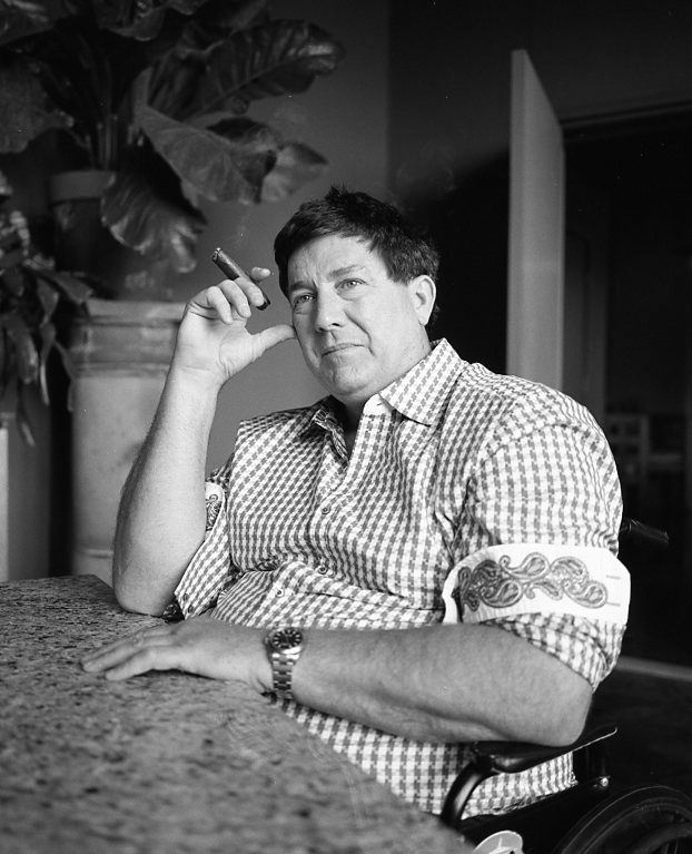 Man seated at a table, holding a cigarette, in a black-and-white indoor portrait. Shot on medium format film by Madison Avery Somerville. Developed and printed in a traditional wet darkroom by the photographer.