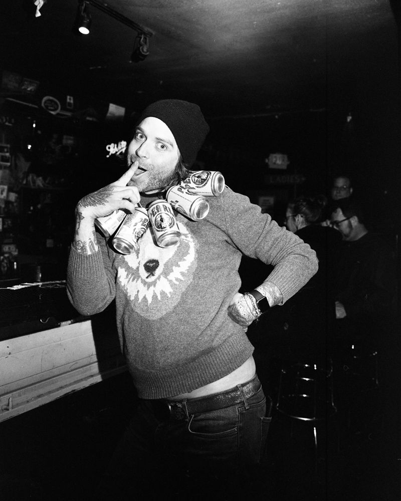 Person posing in a dim bar, wearing a can of pearl beer cans as a necklace, with his hand on hip and looking sideways. Shot on medium format film. Developed and printed in a traditional wet darkroom by the photographer, Madison Avery Somerville.