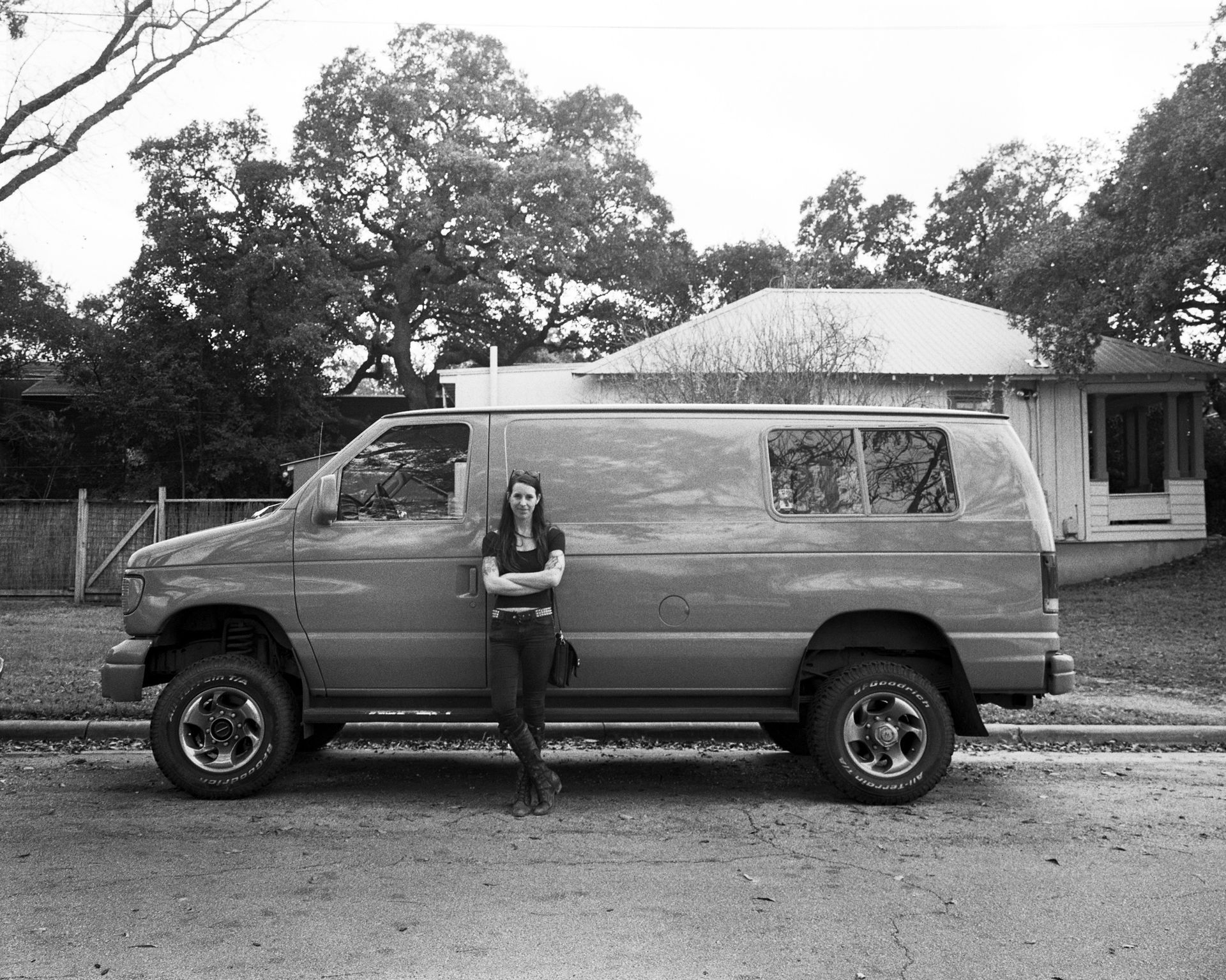 Woman leaning against a van parked in front of a house and trees.Shot on medium format film by Madison Avery Somerville. Developed and printed in a traditional wet darkroom by the photographer.