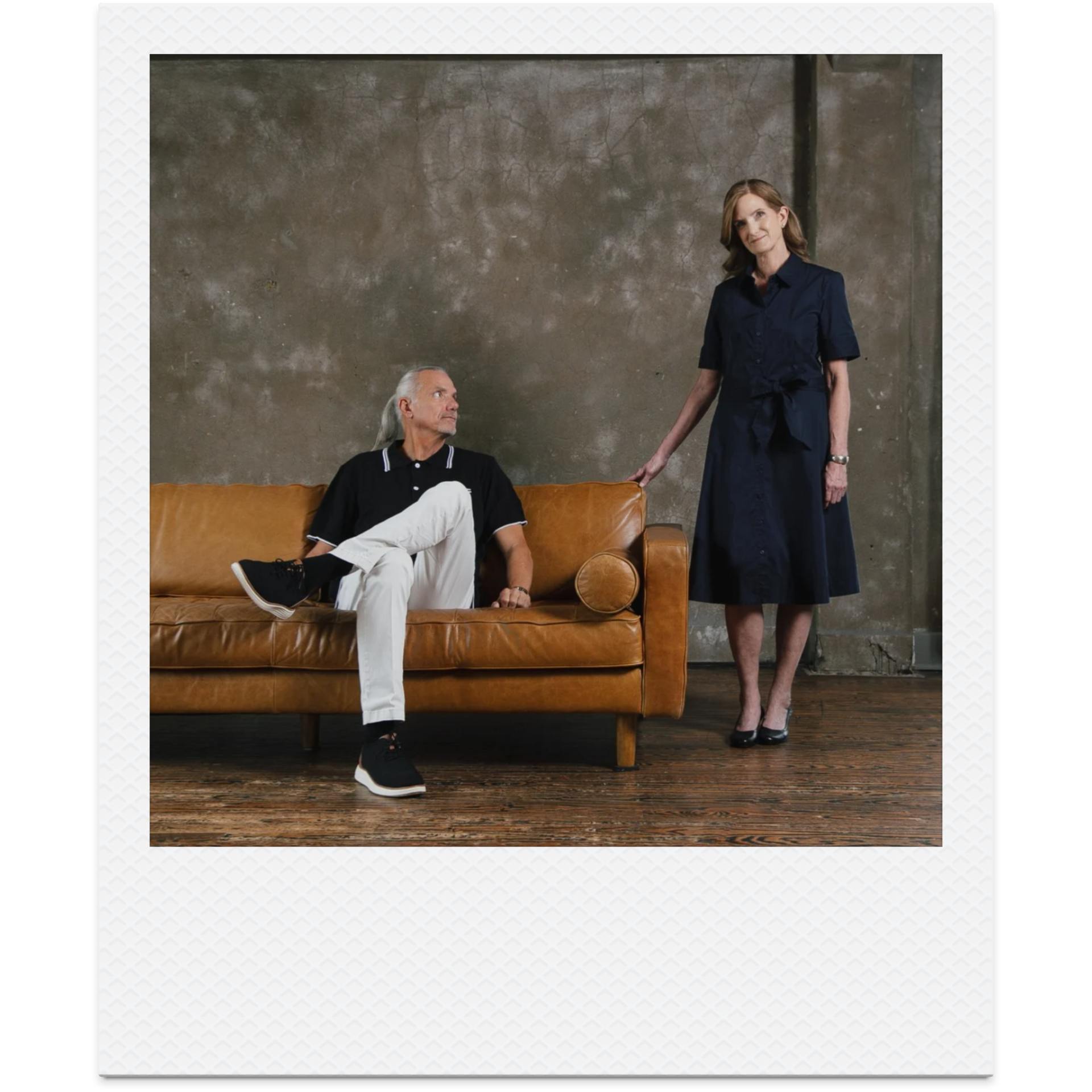A man sits on a brown leather couch and looks up at a woman standing next to it in a blue dress.