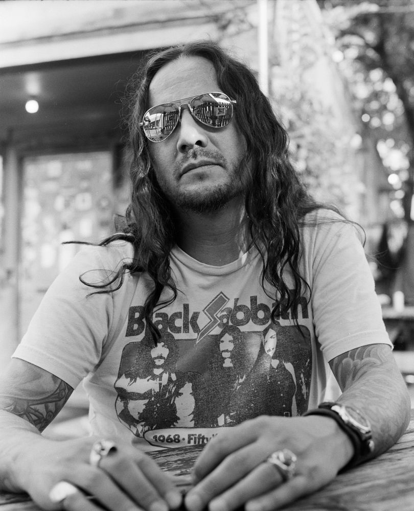 Person with long hair and sunglasses wearing a graphic T-shirt, seated outdoors. Shot on medium format film. Developed and printed in a traditional wet darkroom by the photographer, Madison Avery Somerville.