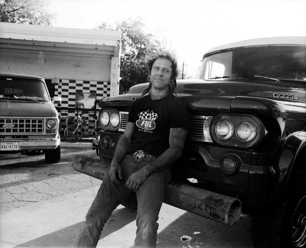 Man seated on a log between two classic trucks in a driveway, black-and-white photo. Shot on medium format film by Madison Avery Somerville. Developed and printed in a traditional wet darkroom by the photographer.