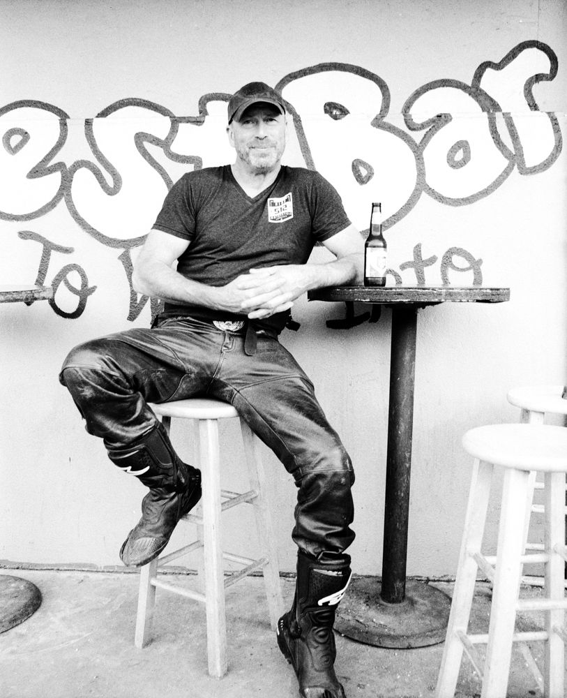 Man sitting on a stool beside a table in outside a bar. Shot on medium format film by Madison Avery Somerville. Developed and printed in a traditional wet darkroom by the photographer.