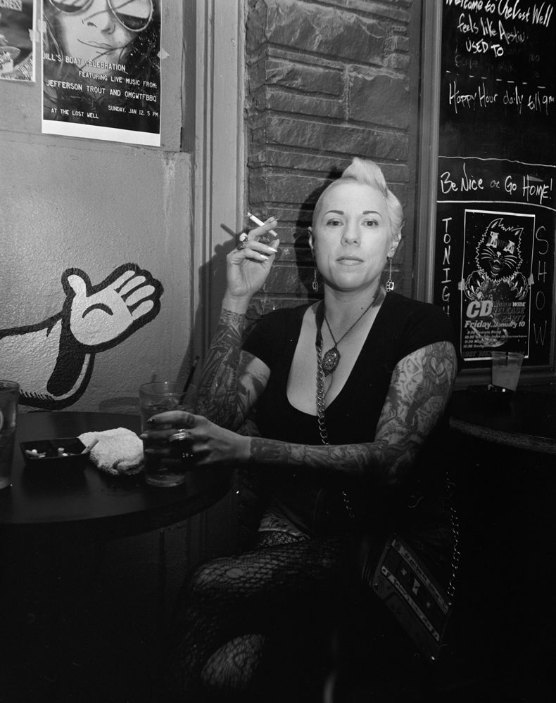 Person with tattoos sits at a table in a dim café, holding a cigarette and looking at the camera. Shot on medium format film by Madison Avery Somerville. Developed and printed in a traditional wet darkroom by the photographer.