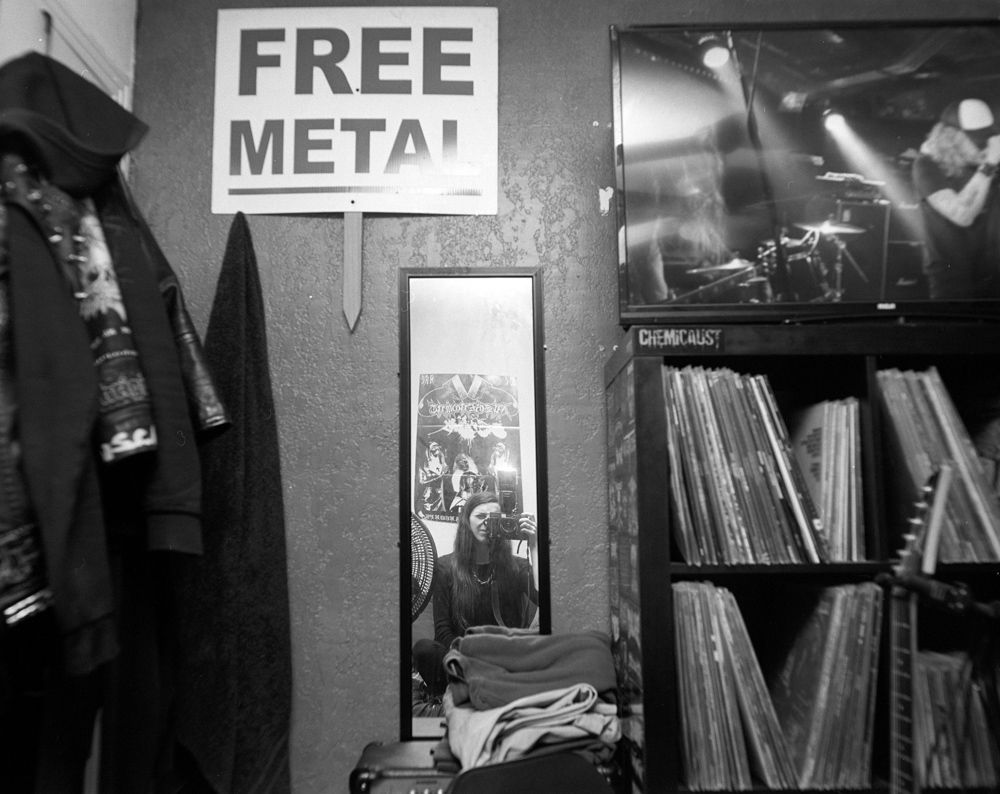 Black-and-white music shop corner with “FREE METAL” sign, hanging clothes, mirror, records, and stage poster. Shot on medium format film by Madison Avery Somerville. Developed and printed in a traditional wet darkroom by the photographer.