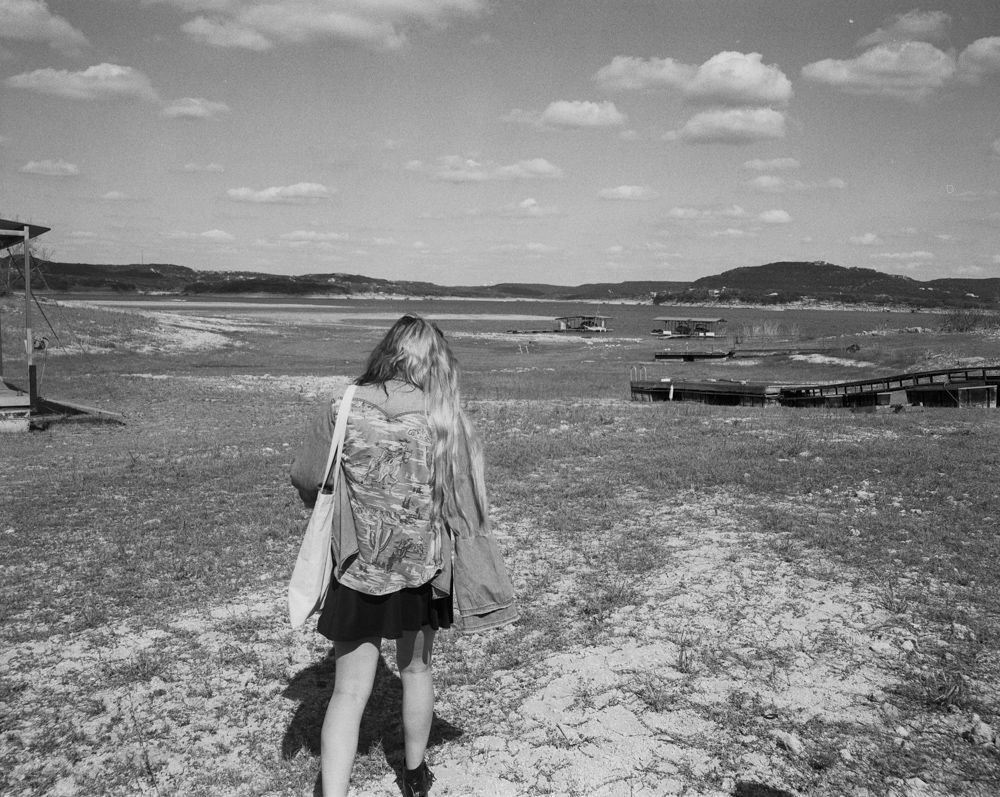 A person with long hair walks across a dry, grassy landscape toward a distant body of water under a cloudy sky.