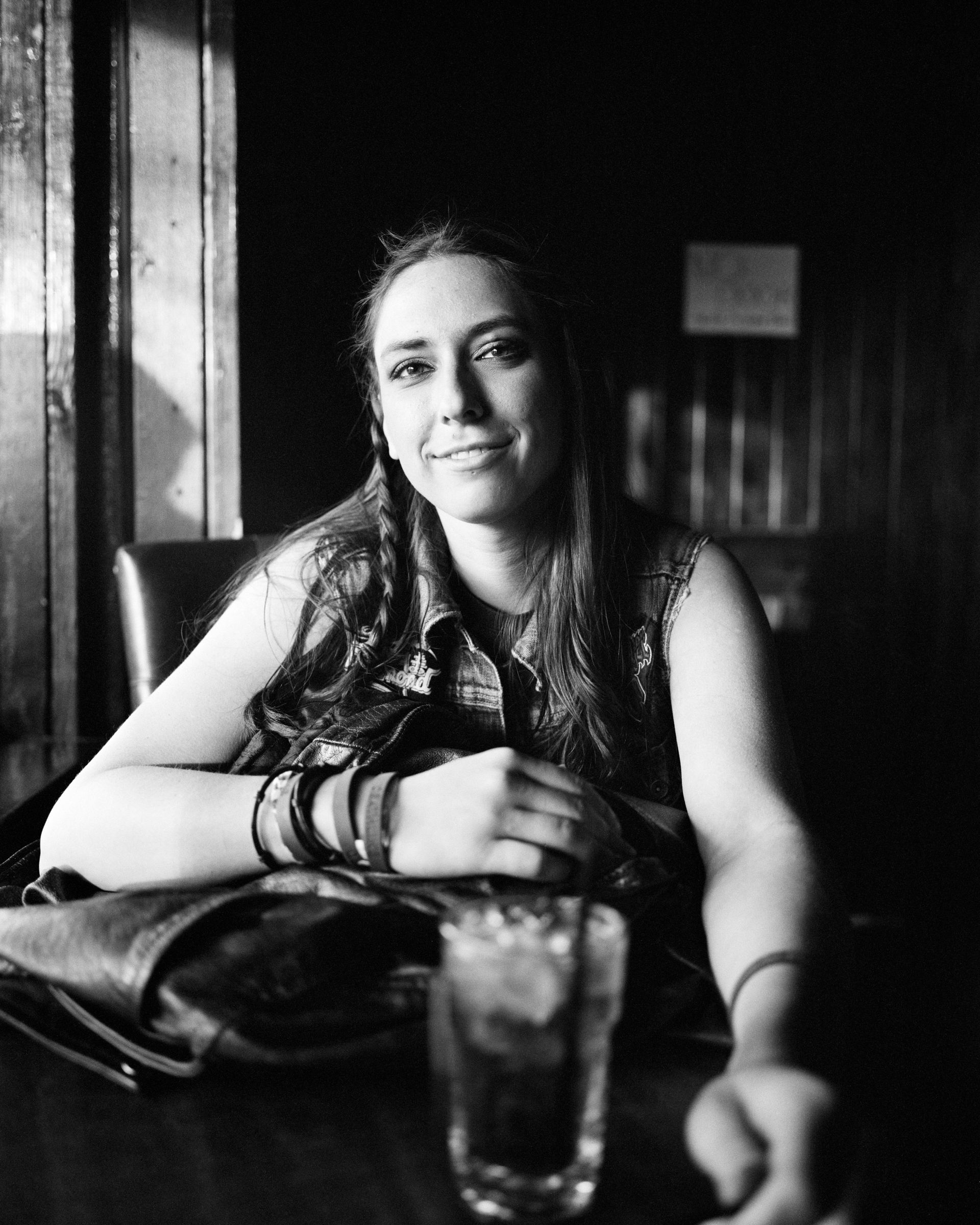 Woman sitting at a table in a dim room, resting her arms near a glass, in black and white.  Shot on medium format film. Developed and printed in a traditional wet darkroom by the photographer, Madison Avery Somerville.