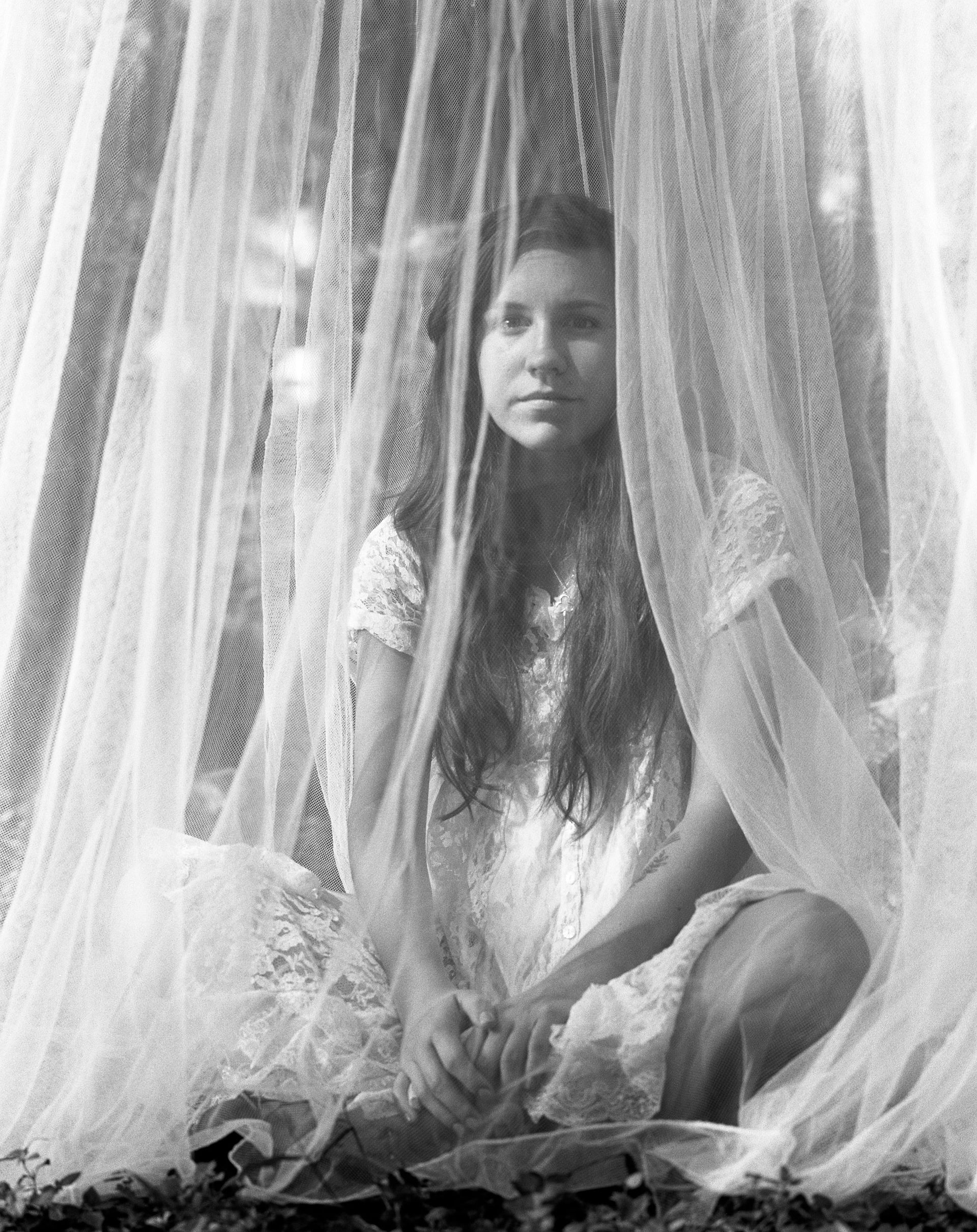 Young person sits behind sheer white curtains in a black-and-white portrait, facing forward. Shot on medium format film by Madison Avery Somerville. Developed and printed in a traditional wet darkroom by the photographer.