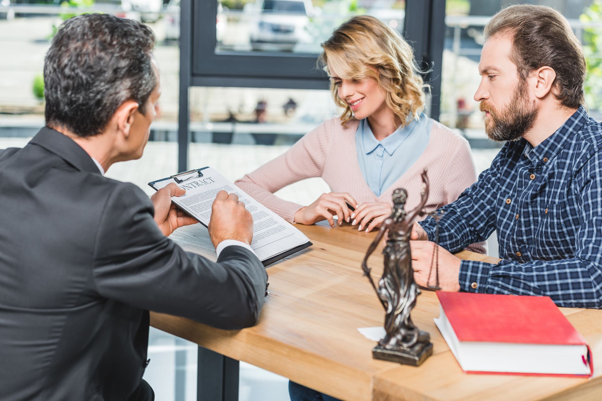An attorney reviewing a contract with a smiling couple at a desk, with a Lady Justice statue nearby