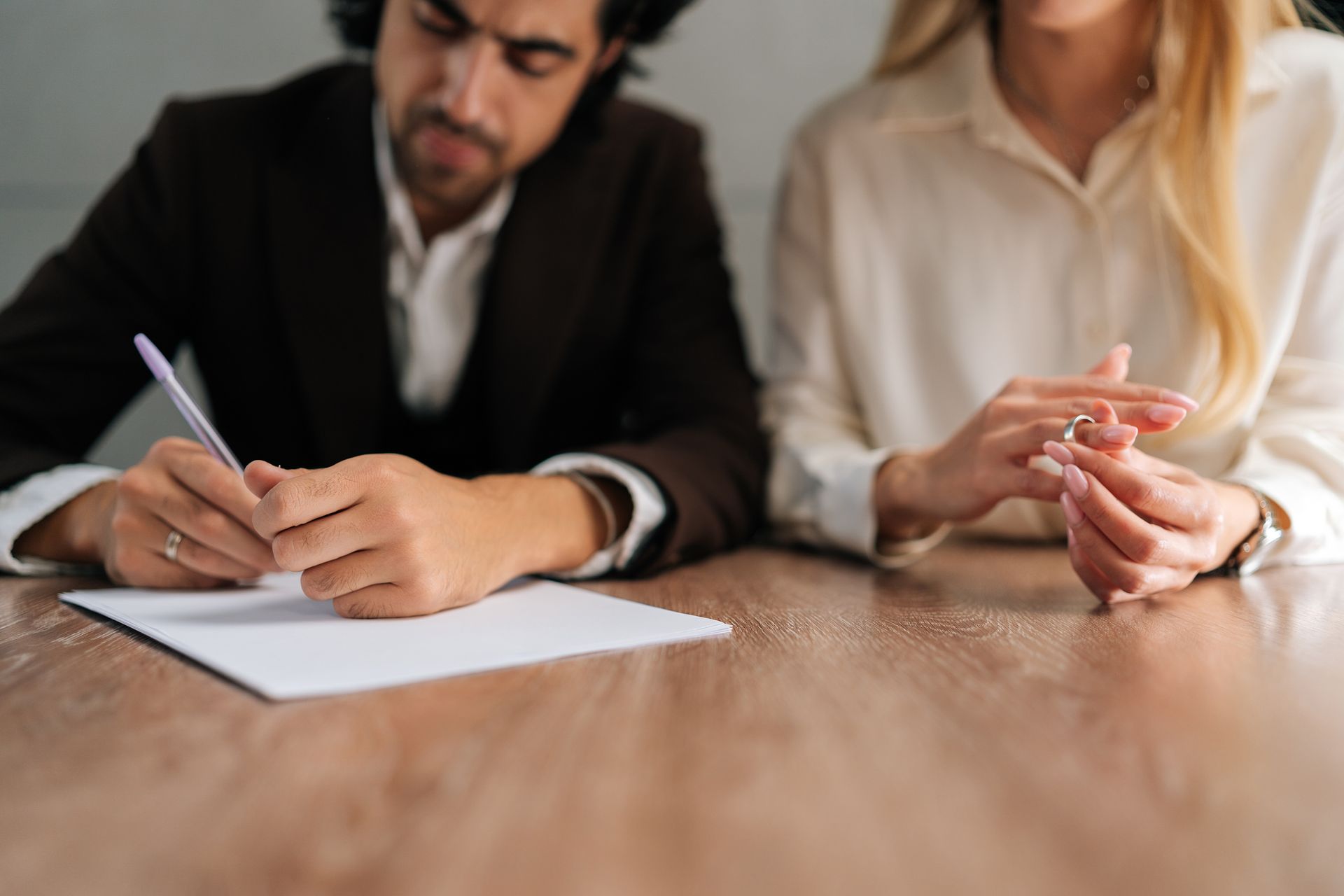 Man in suit signing divorce paperwork while woman removes her wedding ring at a wooden table Man in suit signing divorce paperwork while woman removes her wedding ring at a wooden table