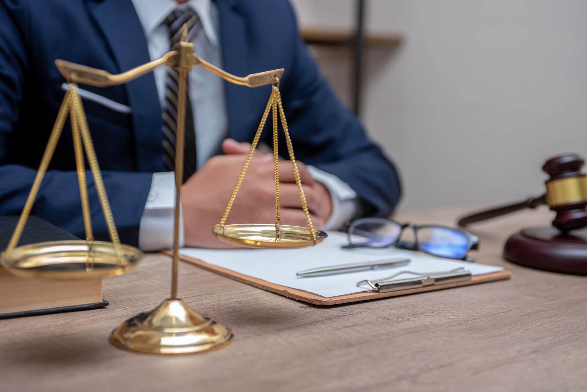 Scales of justice, legal documents, and gavel on a desk with a person in a suit, suggesting legal counsel. Scales of justice, legal documents, and gavel on a desk with a person in a suit, suggesting legal counsel.