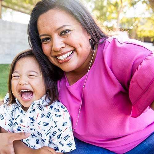 A mom and young child are sitting outside at a park and smiling.