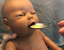 A baby doll is being fed with a spoon by a hand, with its mouth open.