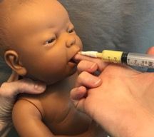 Hand feeding a baby doll with a syringe-like tube and a caregiver’s hands visible.