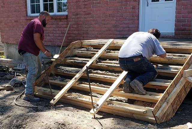 Deux hommes travaillent sur un escalier en bois devant un immeuble en brique.