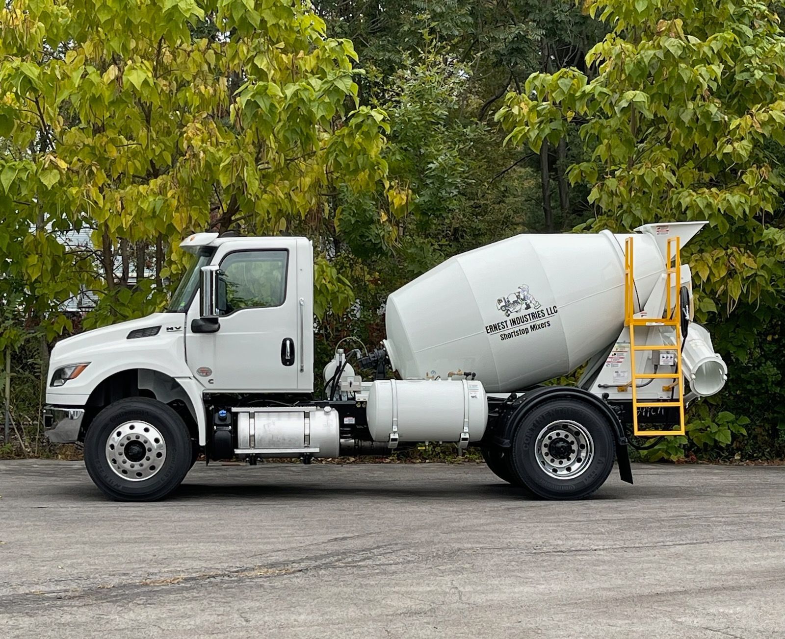 Camion malaxeur à béton blanc garé à l'extérieur avec feuillage d'automne en arrière-plan.