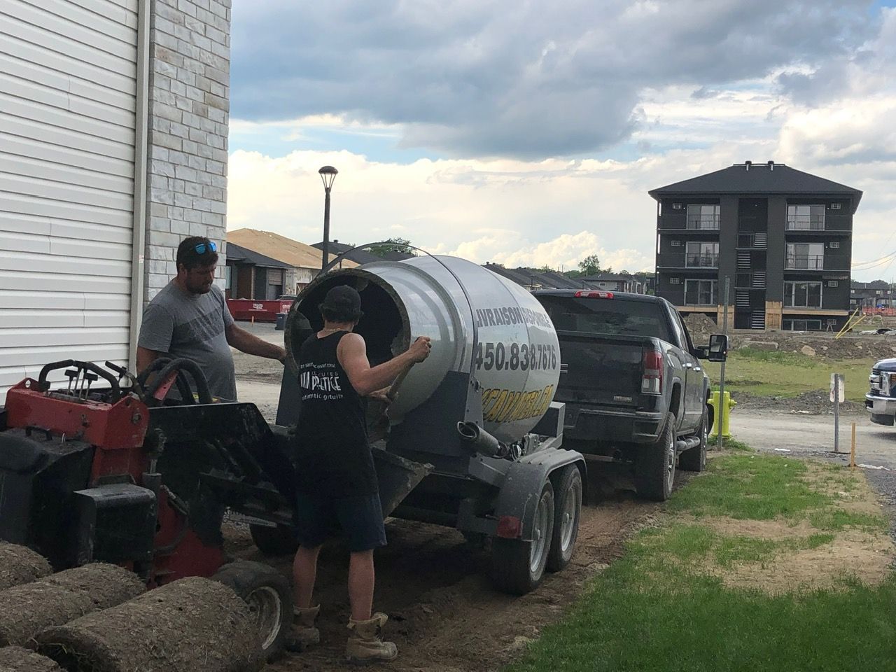 Deux hommes travaillant avec une bétonnière et un camion, à côté d'un bâtiment et de quelques rouleaux de gazon.