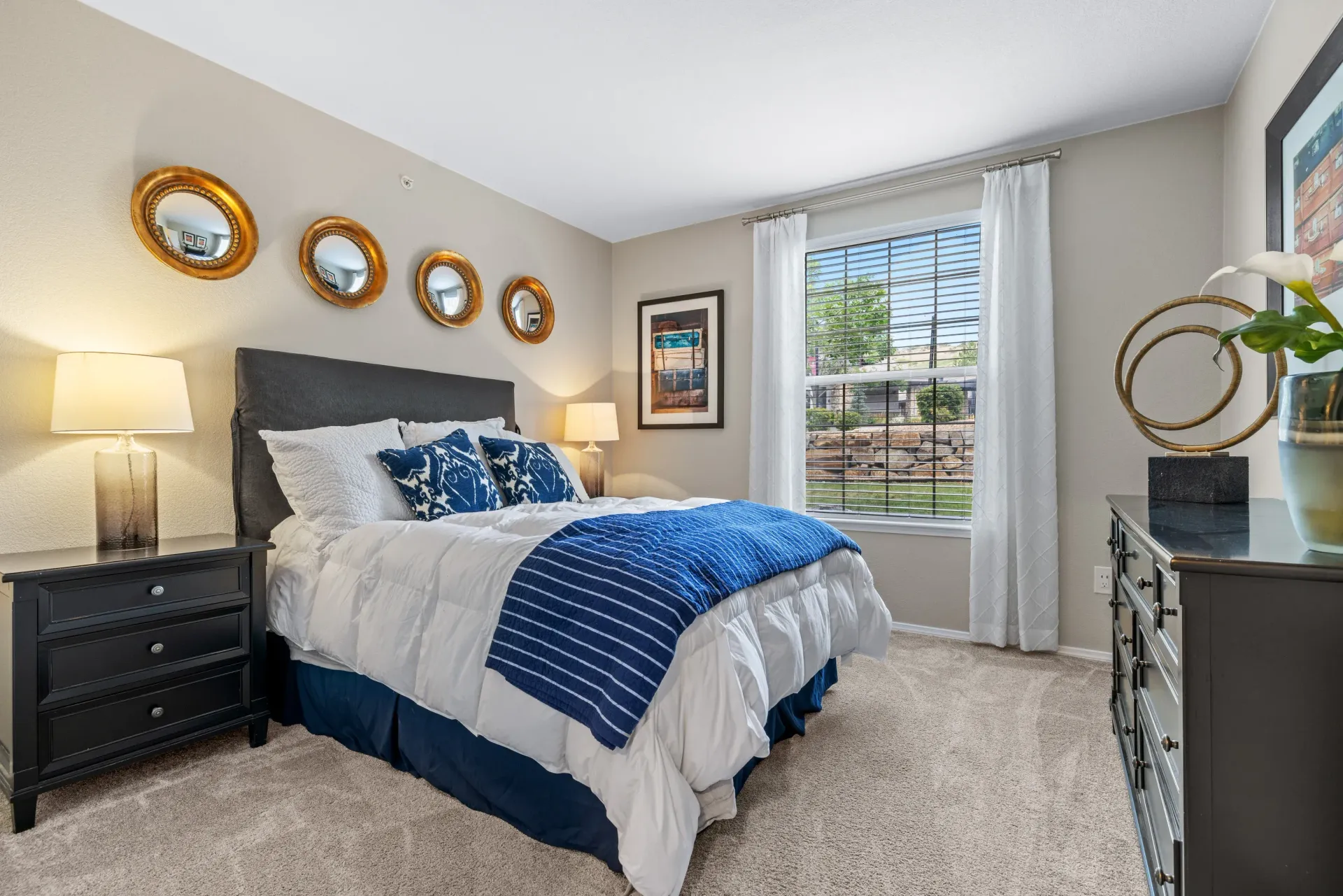 Furnished bedroom with a bed, nightstands, dresser, and a window with white curtains at Montrachet apartments in Lakewood, CO.
