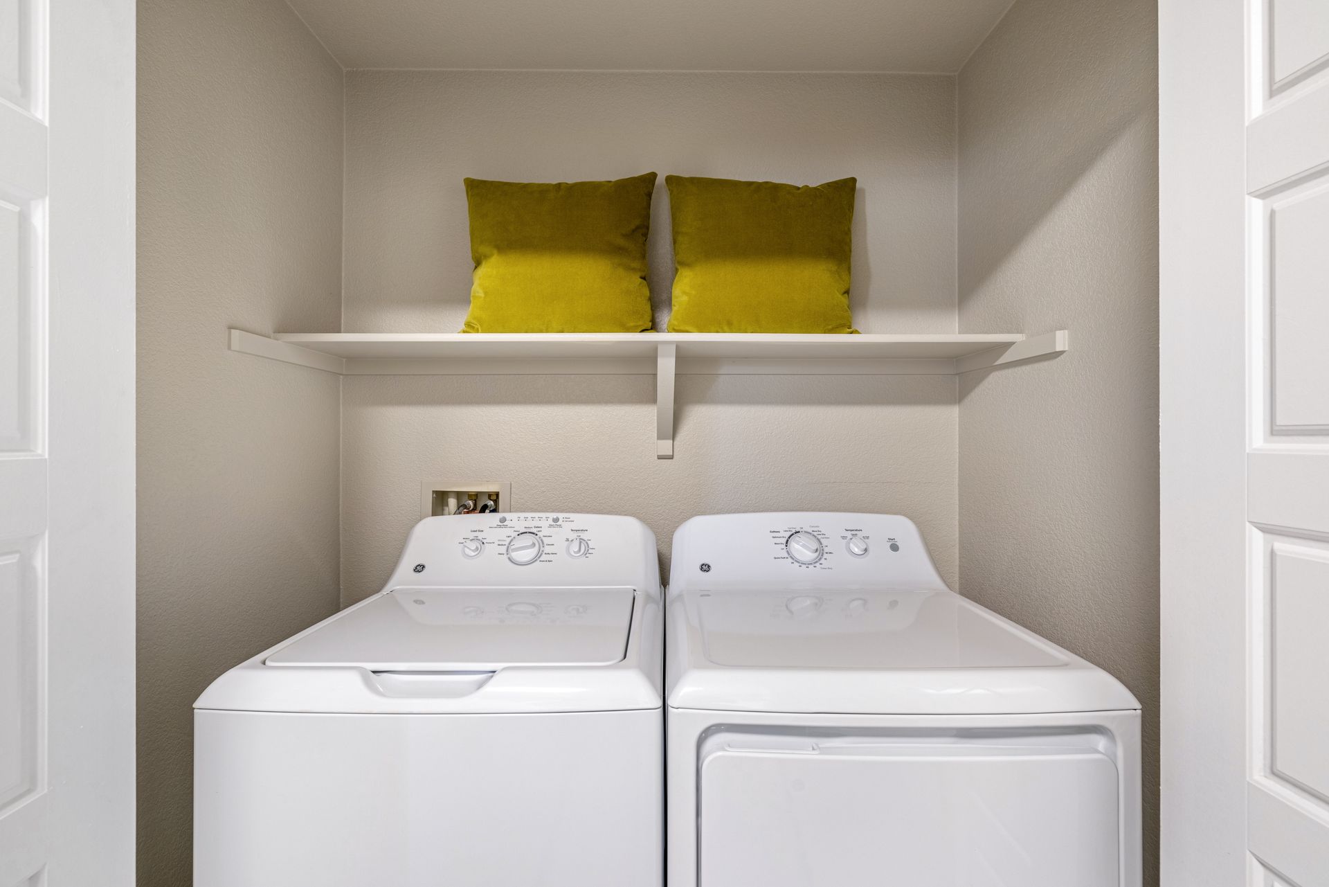 Laundry room with white washer and dryer, shelf with two yellow pillows at Montrachet apartments in Lakewood, CO.