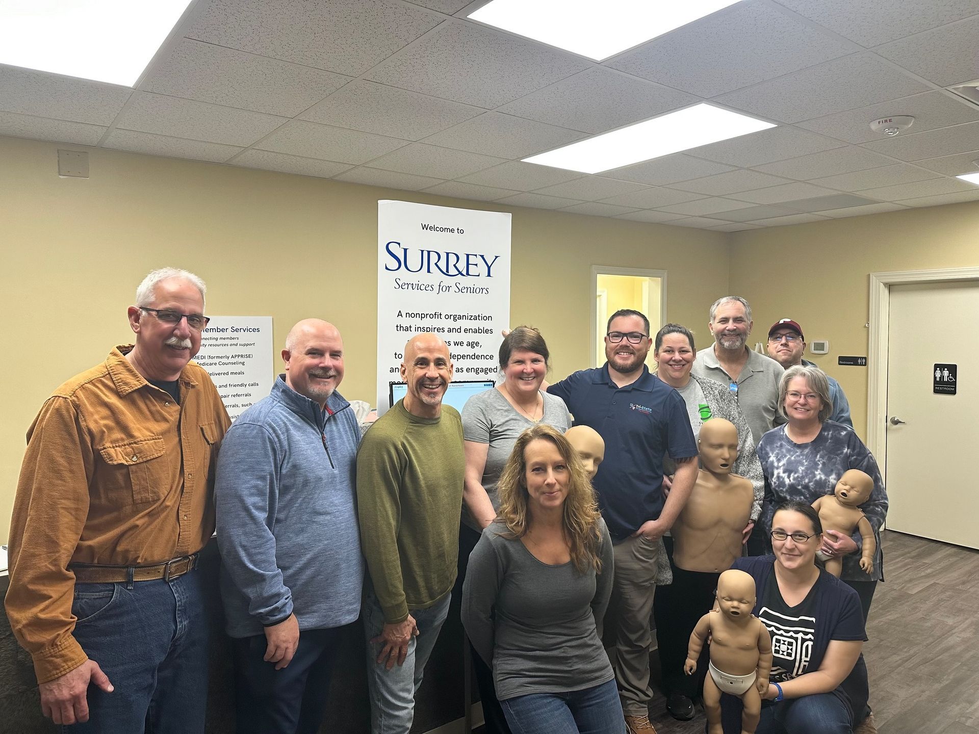 Group of people posing indoors in a beige room, smiling near a sign that says “Surrey.”