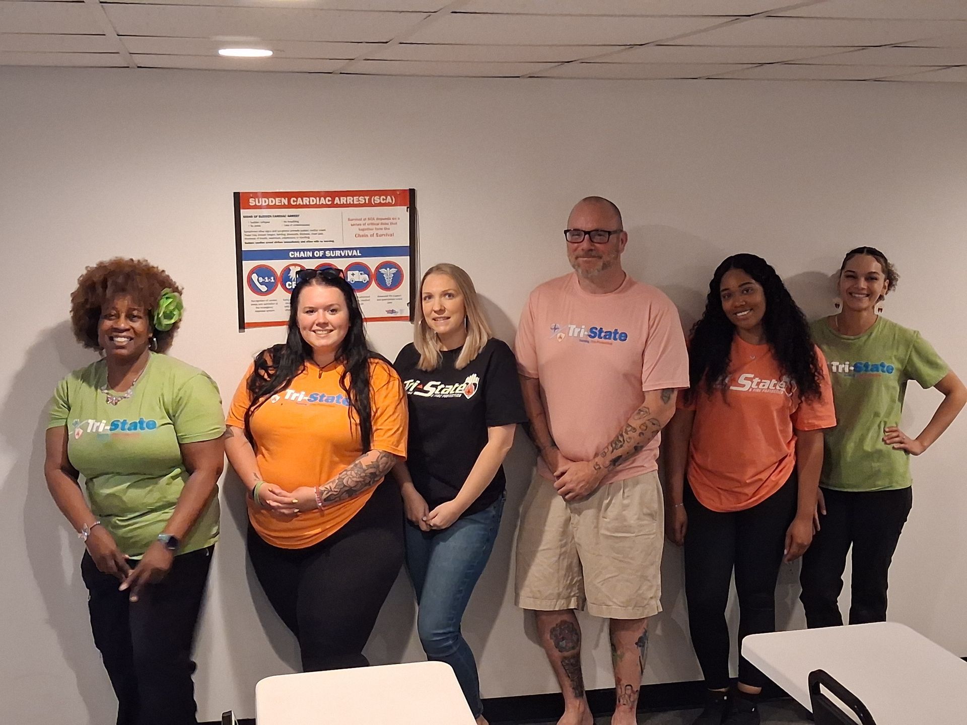 Six adults stand in a row indoors, smiling in casual colorful shirts before a wall sign.